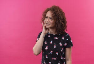 young woman with short curly hair looking unwell touching cheek having toothache standing pink wall